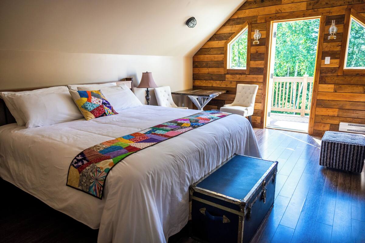 A bedroom with a large bed, a quilt, a desk, a chair, and a view of the outdoors.