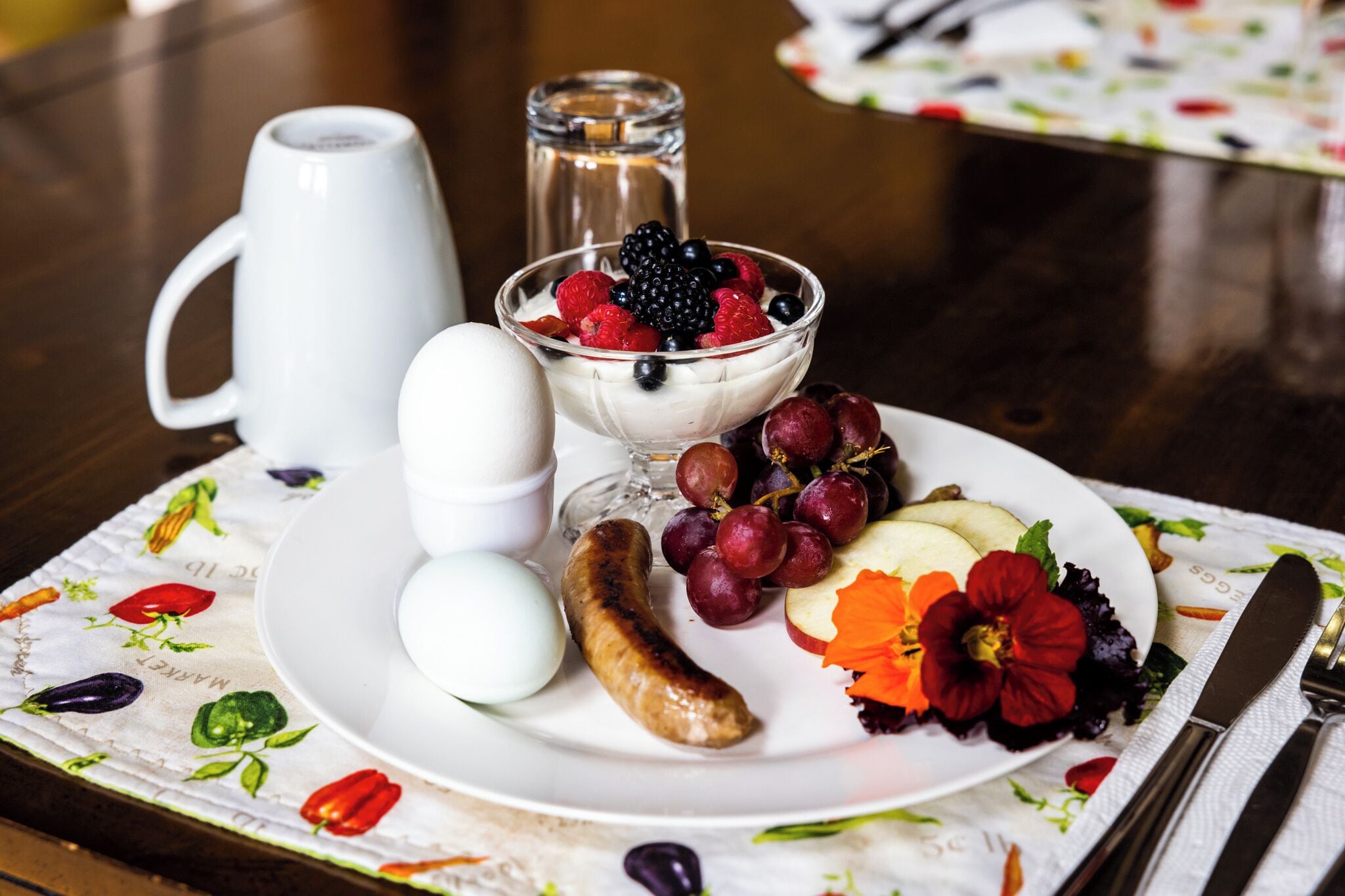 Un plato de desayuno con huevos, salchicha, uvas, bayas y una flor.