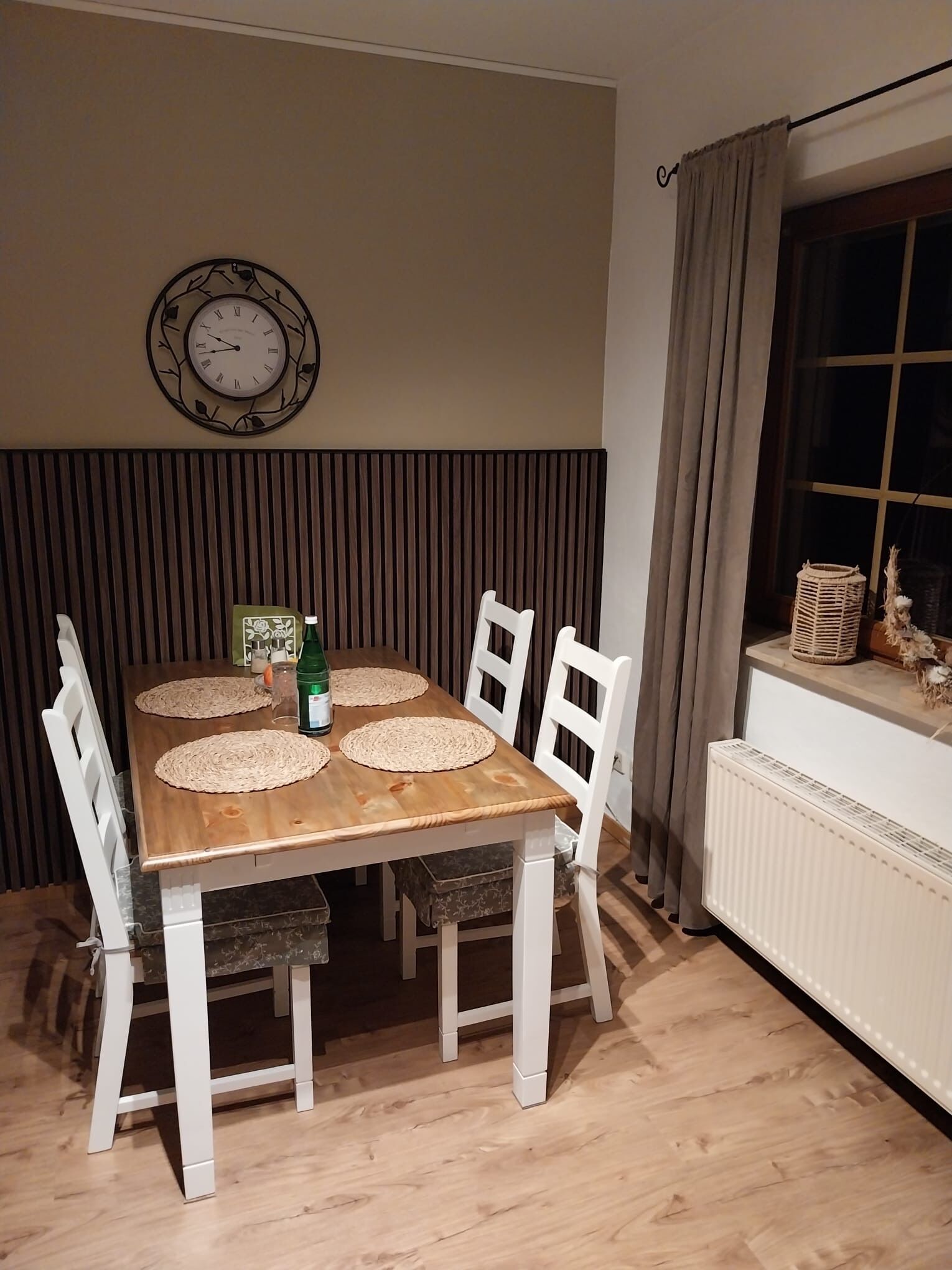 A dining area with a wooden table, white chairs, a wall-mounted clock, and a window with curtains.