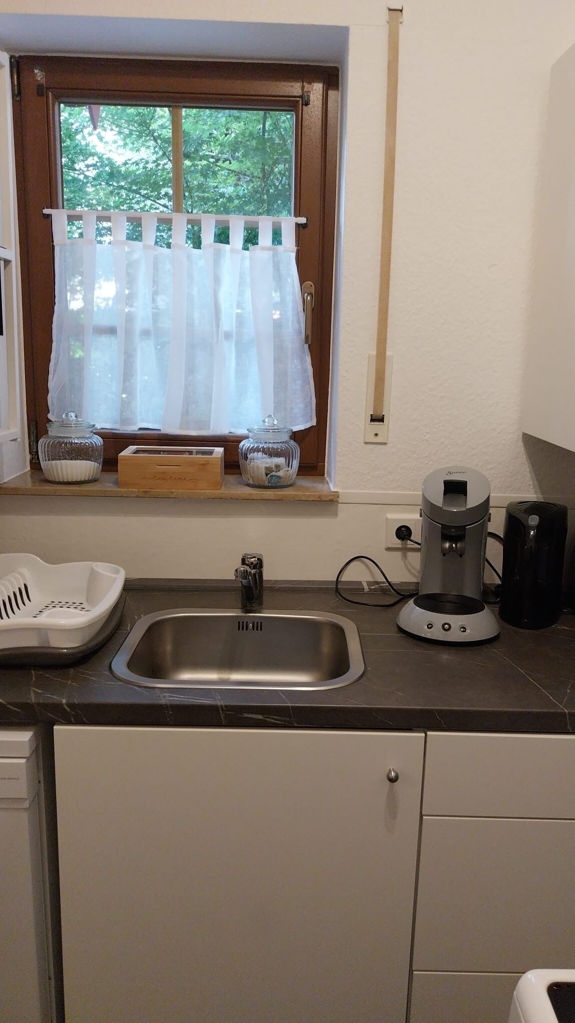 A kitchen with a stainless steel sink, a coffee maker, and a window with a white curtain.