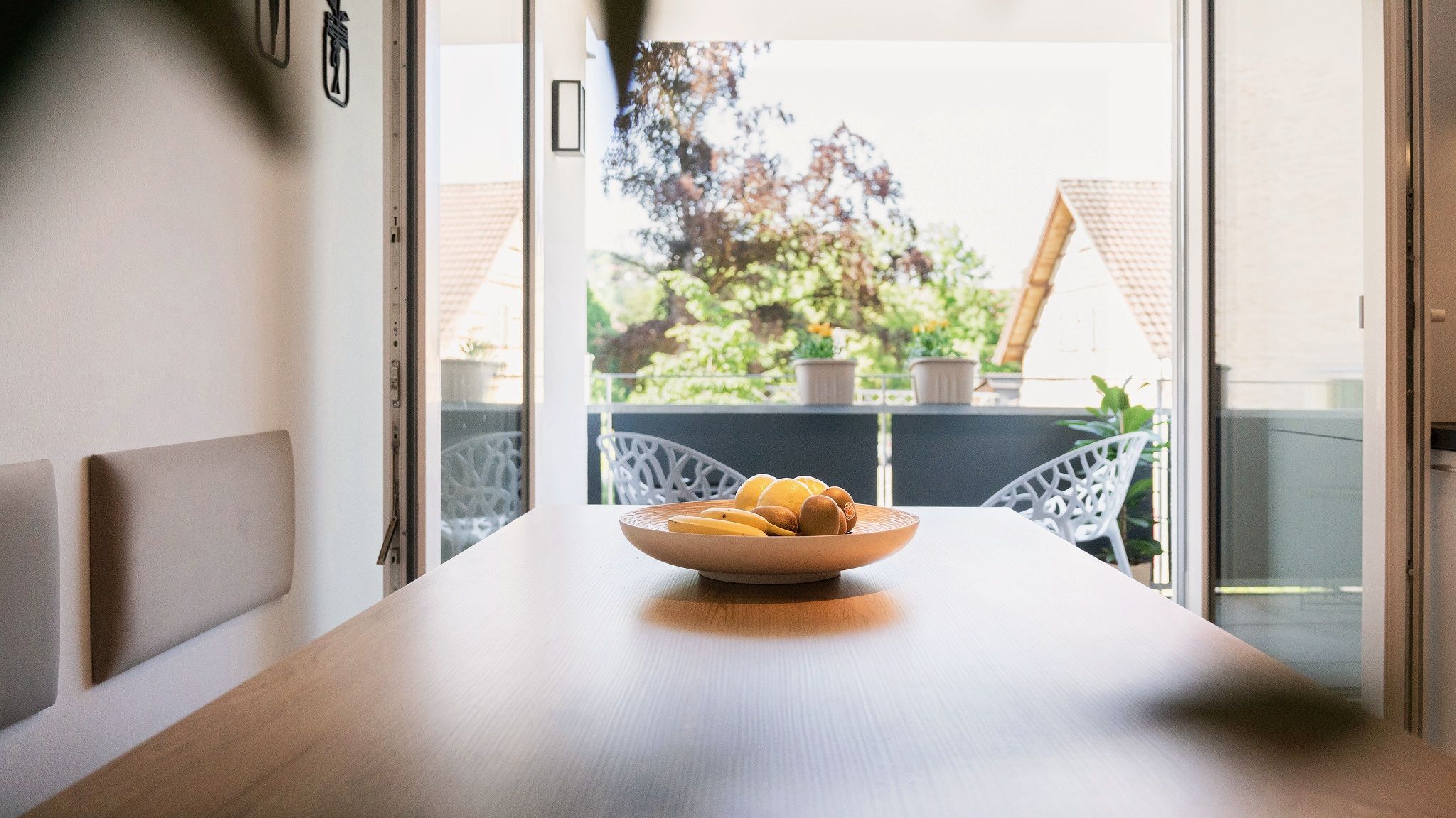 A dining table with a bowl of fruit in the center, white chairs, and a view of a balcony with plants and a house in the background.