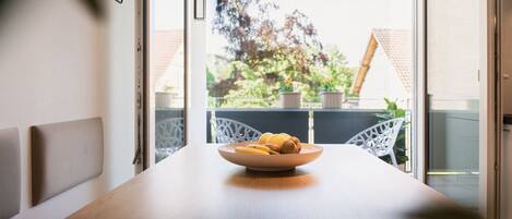 A dining table with a bowl of fruit in the center, white chairs, and a view of a balcony with plants and a house in the background.