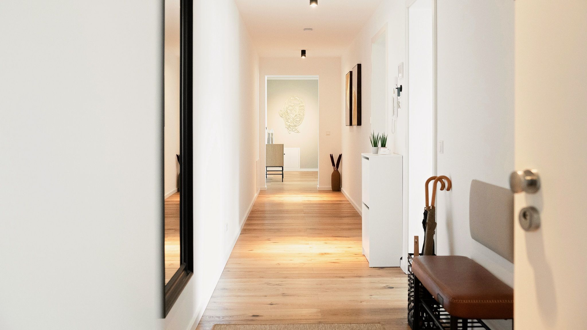 Un pasillo con suelo de madera, un banco, una puerta con perchero y una planta en la pared.