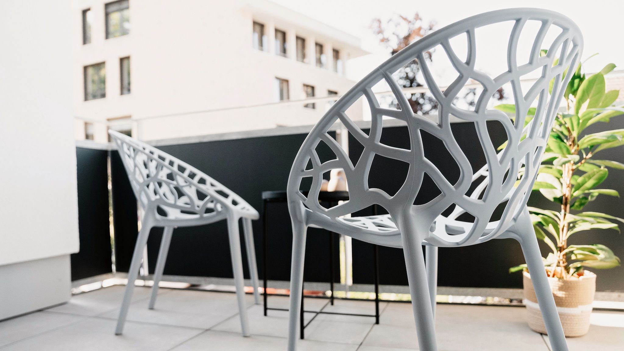 Zwei weiße Metallstühle mit geometrischem Muster stehen auf einem Balkon mit einem Blumentopf.