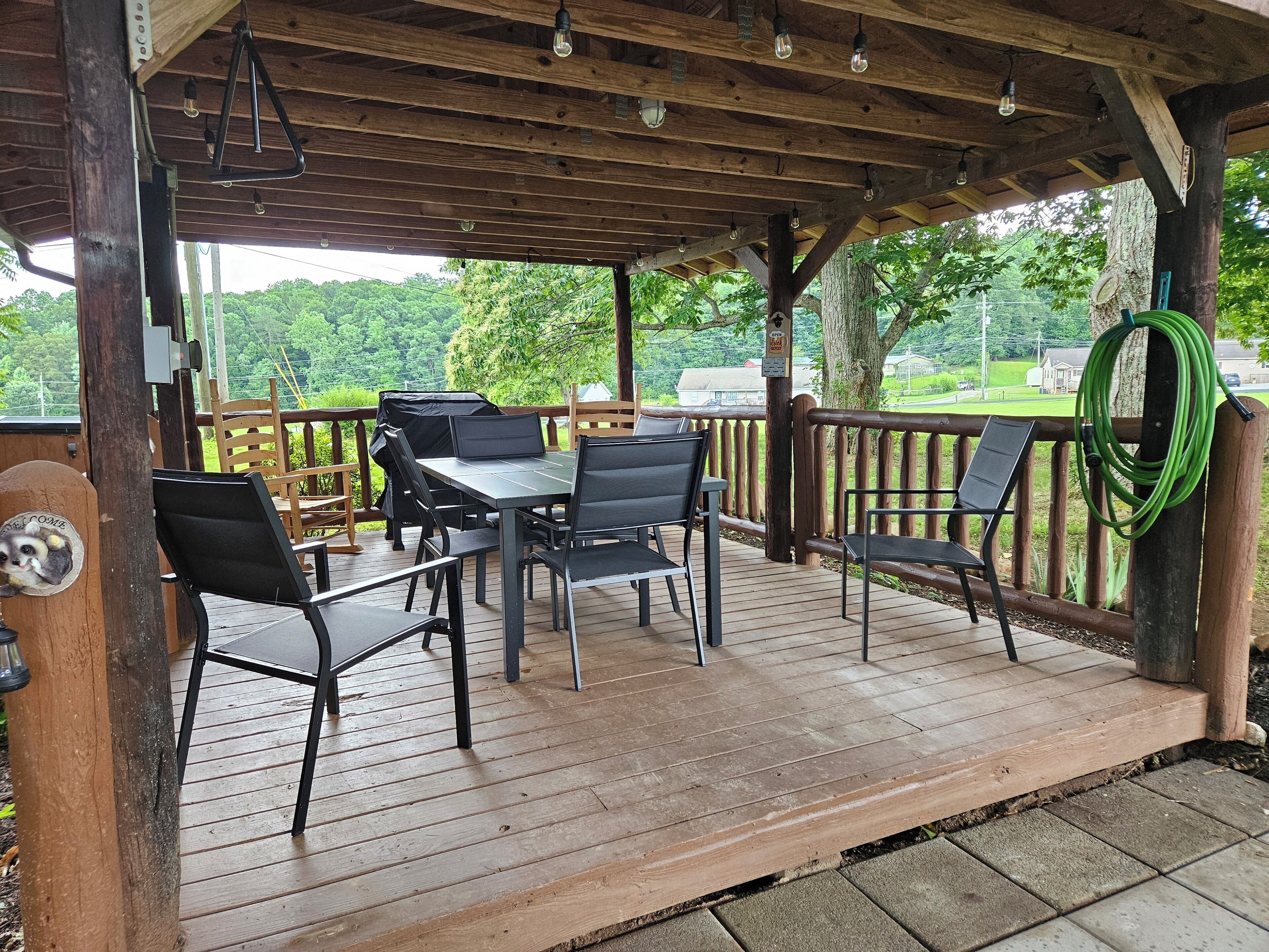 Una veranda esterna coperta con tavoli e sedie in legno, un barbecue e vista su un paesaggio verdeggiante.