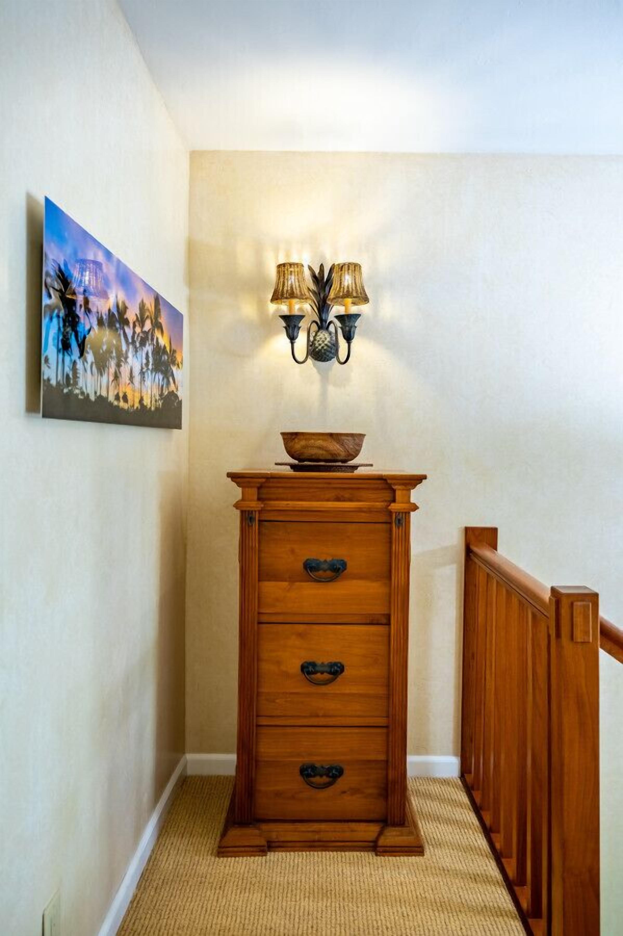 Une commode en bois sur laquelle est posé un bol, une lampe fixée au mur et un tableau accroché au mur.