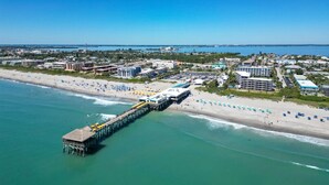 An aerial view of a coastal area with a pier extending into the water, a beach with umbrellas, and buildings in the background. - HARDING HAVEN Spacious pool home 1 block to the beach, walk to Cocoa Beach Pier! (Cocoa Beach)