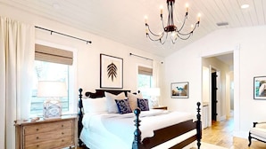 A bedroom with a large bed, wooden headboard, a chandelier, a dresser, and a large window with curtains. - HARDING HAVEN Spacious pool home 1 block to the beach, walk to Cocoa Beach Pier! (Cocoa Beach)