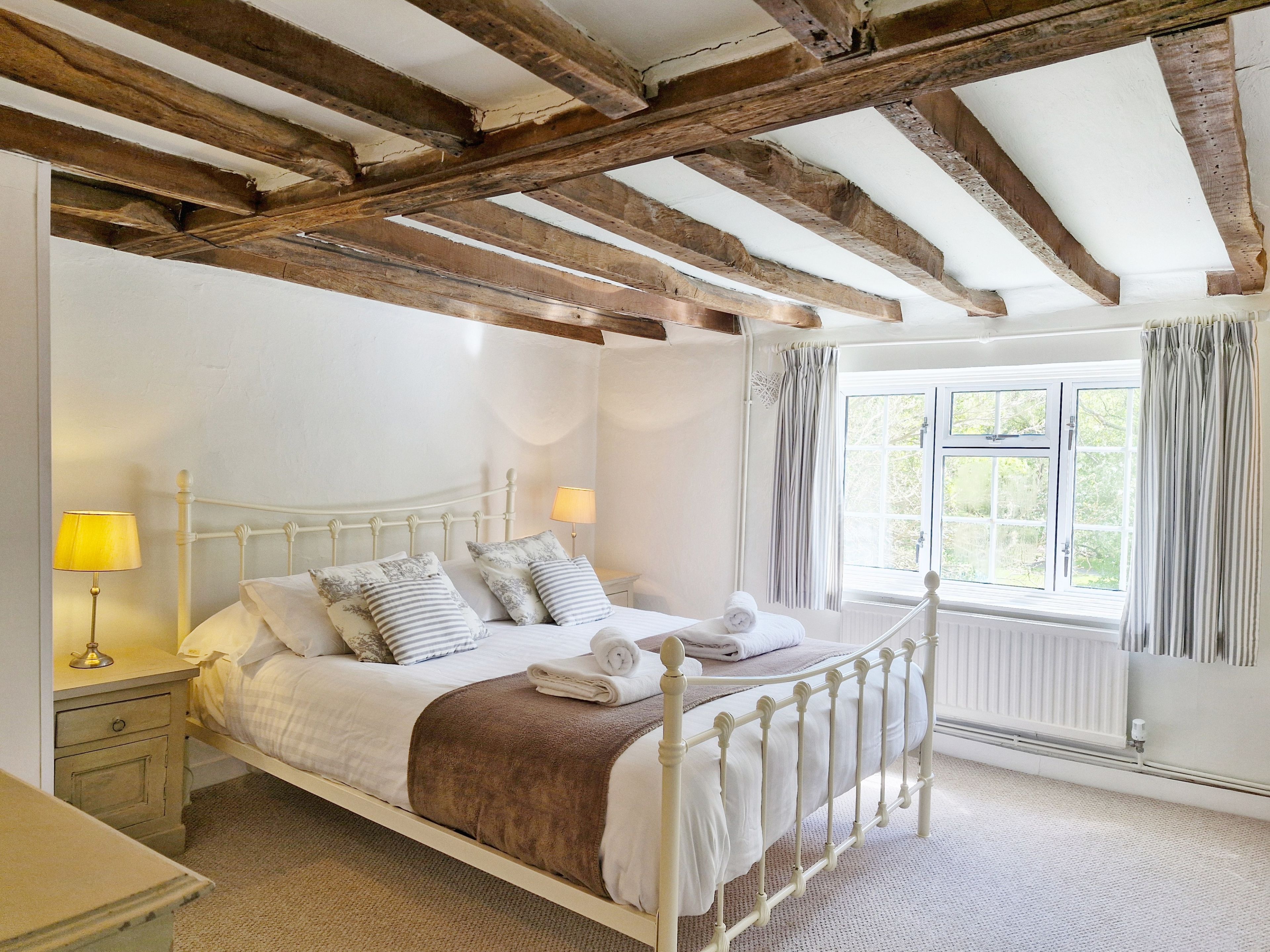 Un dormitorio con estructura de cama metálica blanca, techo de madera, ventana con cortinas y una lámpara de noche.