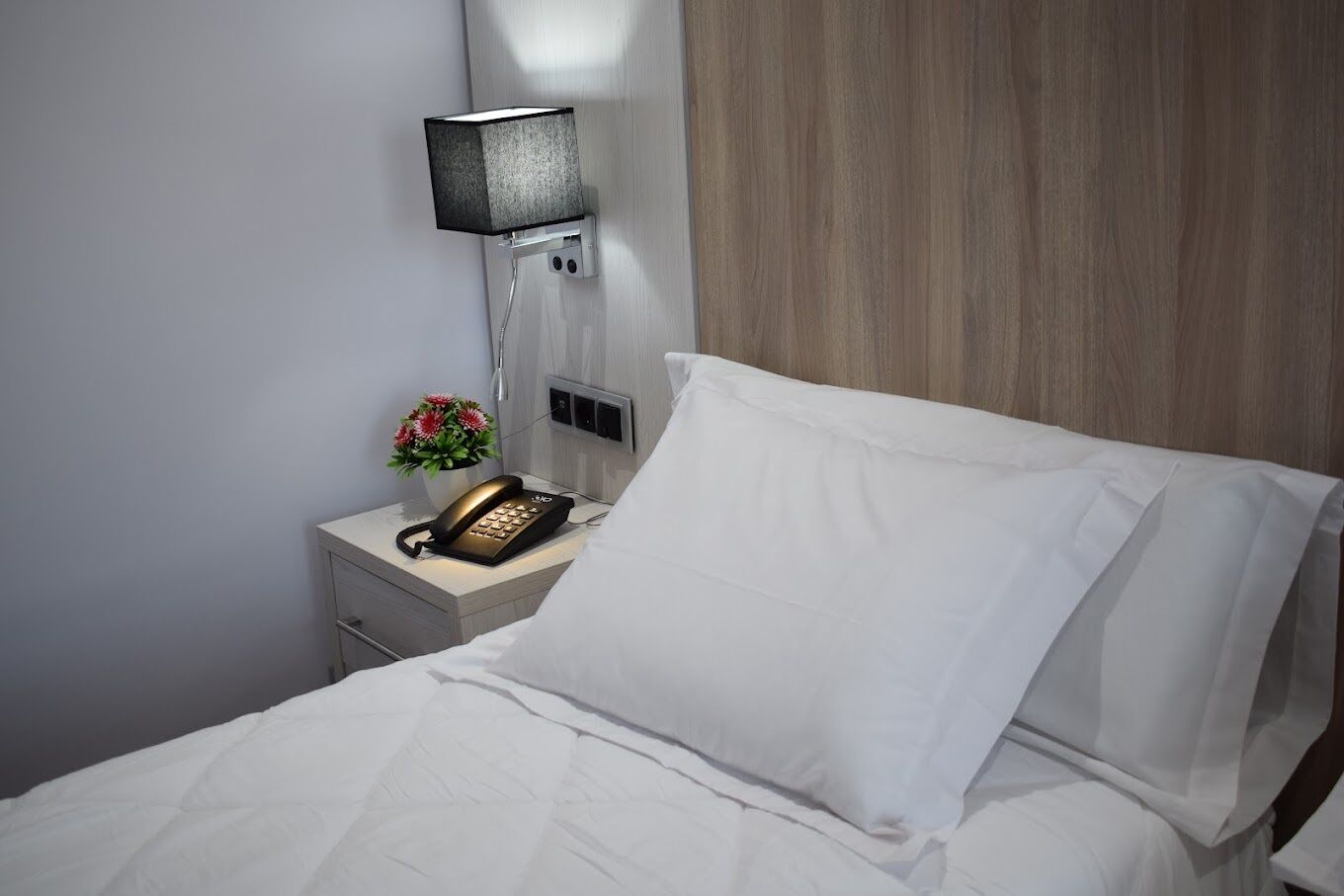 A hotel room with a neatly made bed, a bedside telephone, and a wall-mounted lamp.