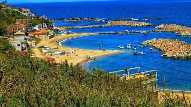 Un pueblo costero con playa, barcos y rompeolas rocosas.