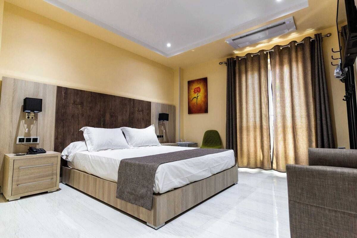 A hotel room with a large bed, bedside tables, a chair, a desk, and an air conditioning unit.