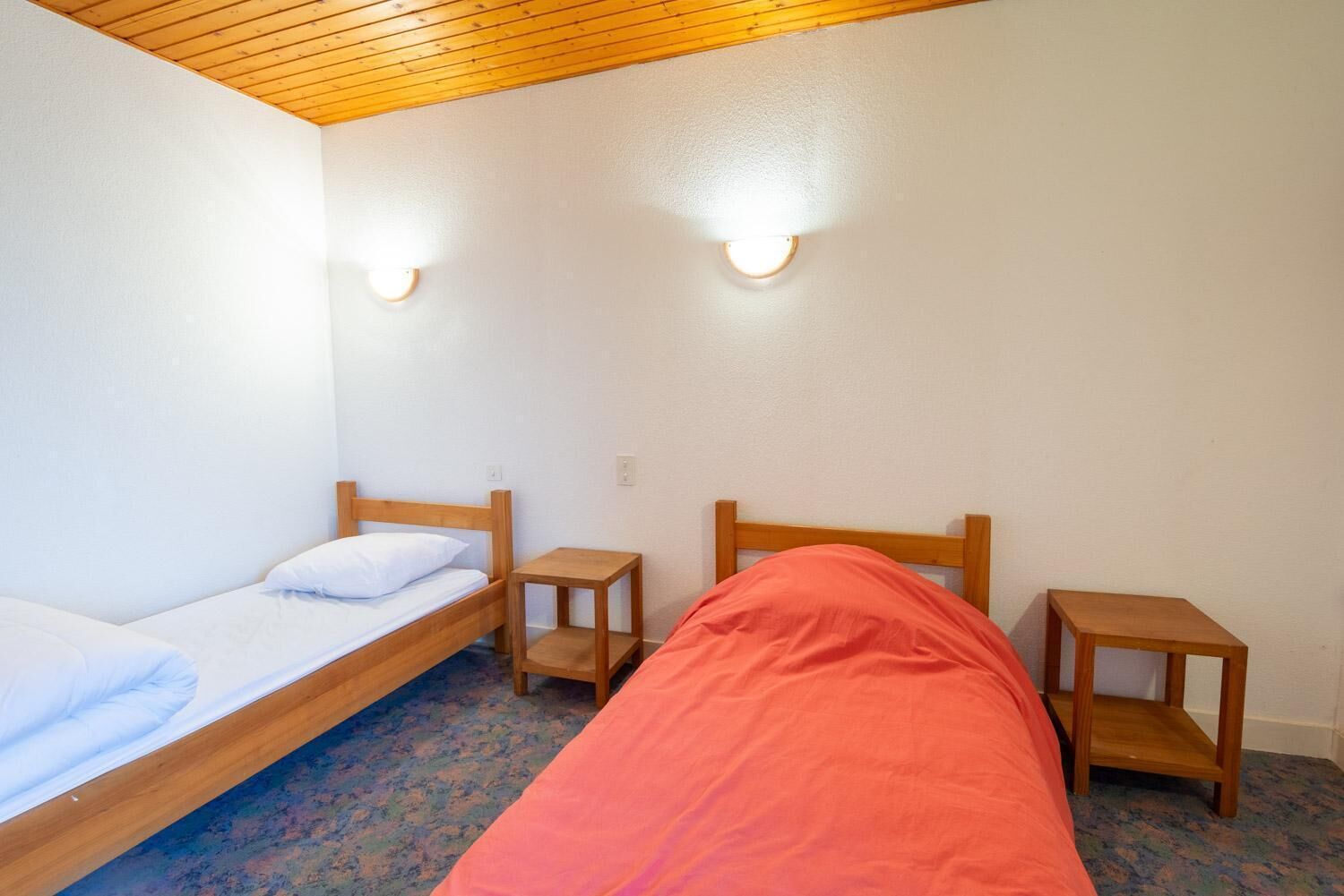 Une petite chambre simple, avec deux lits simples, des têtes de lit en bois et deux tables de chevet.