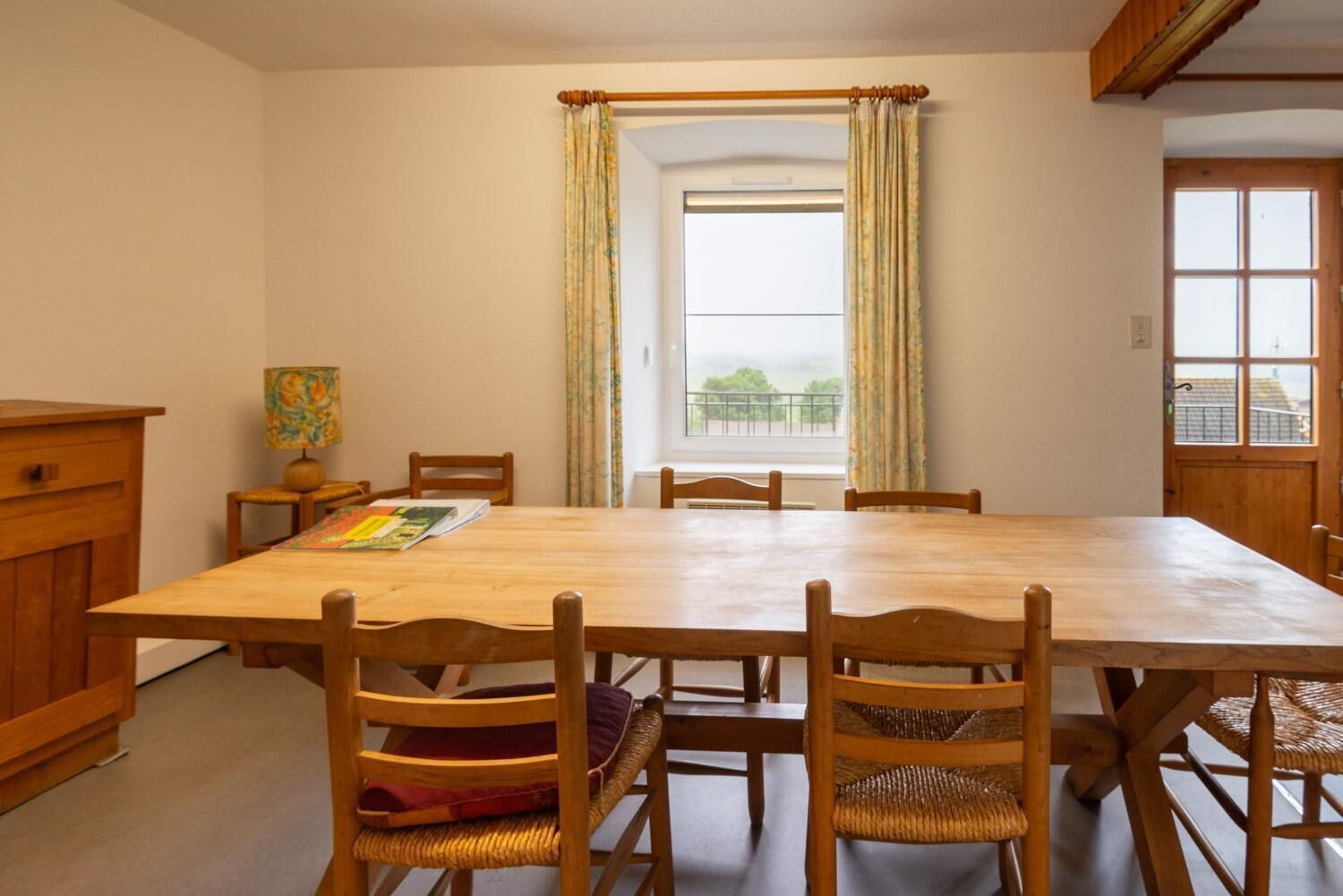 A dining room with a large wooden table, six wooden chairs, a small side table with a lamp, and a window with curtains.