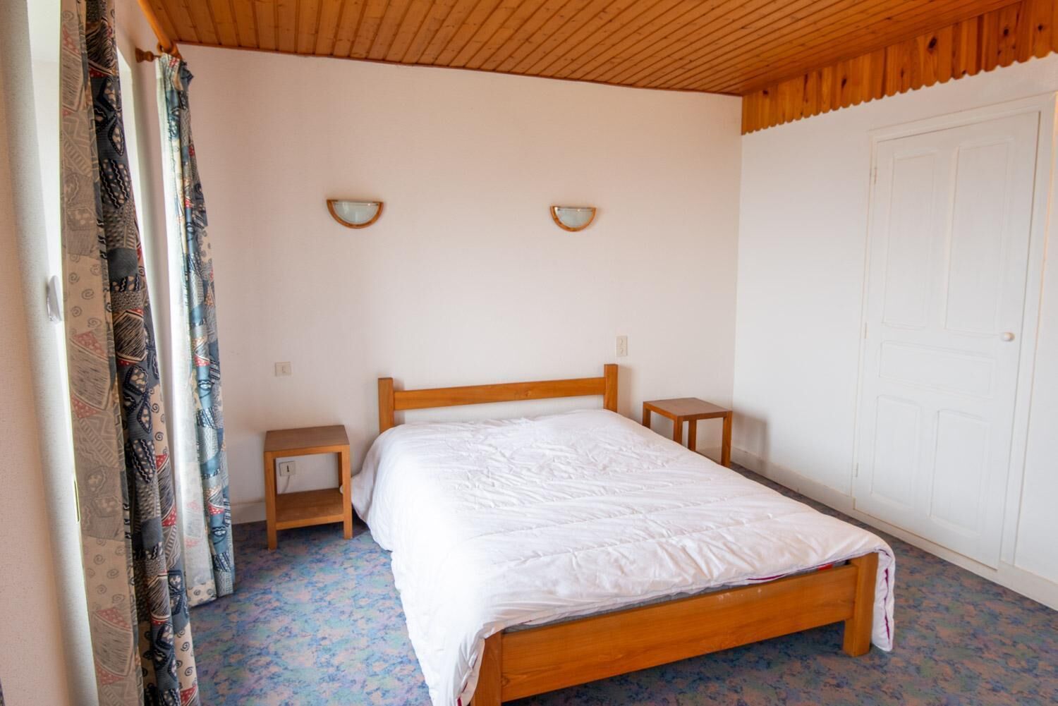 A bedroom with a wooden bed, two bedside tables, patterned curtains, and a white door.