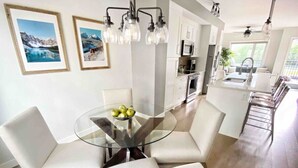 A modern kitchen with white cabinets, a central island with a sink, and a dining area with a glass table and white chairs. - Modern Mountain Townhouse: private garage/entrance (Canmore)