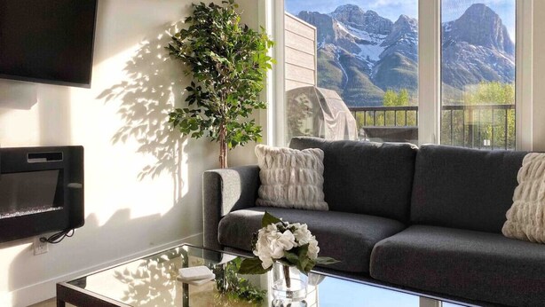 A modern living room with a grey sofa, a glass coffee table, a flat-screen TV, and a large window with a mountain view. - Modern Mountain Townhouse: private garage/entrance (Canmore)