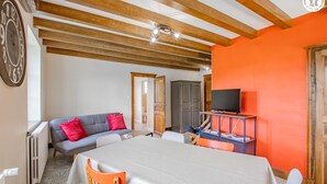 Een kamer met een houten plafond, een grijze bank met rode kussens, een witte tafel en een televisie op een plank.