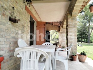 Un patio cubierto con pared de ladrillo, una puerta de madera y una mesa con sillas blancas. Hay plantas en macetas y un colgador de luz.