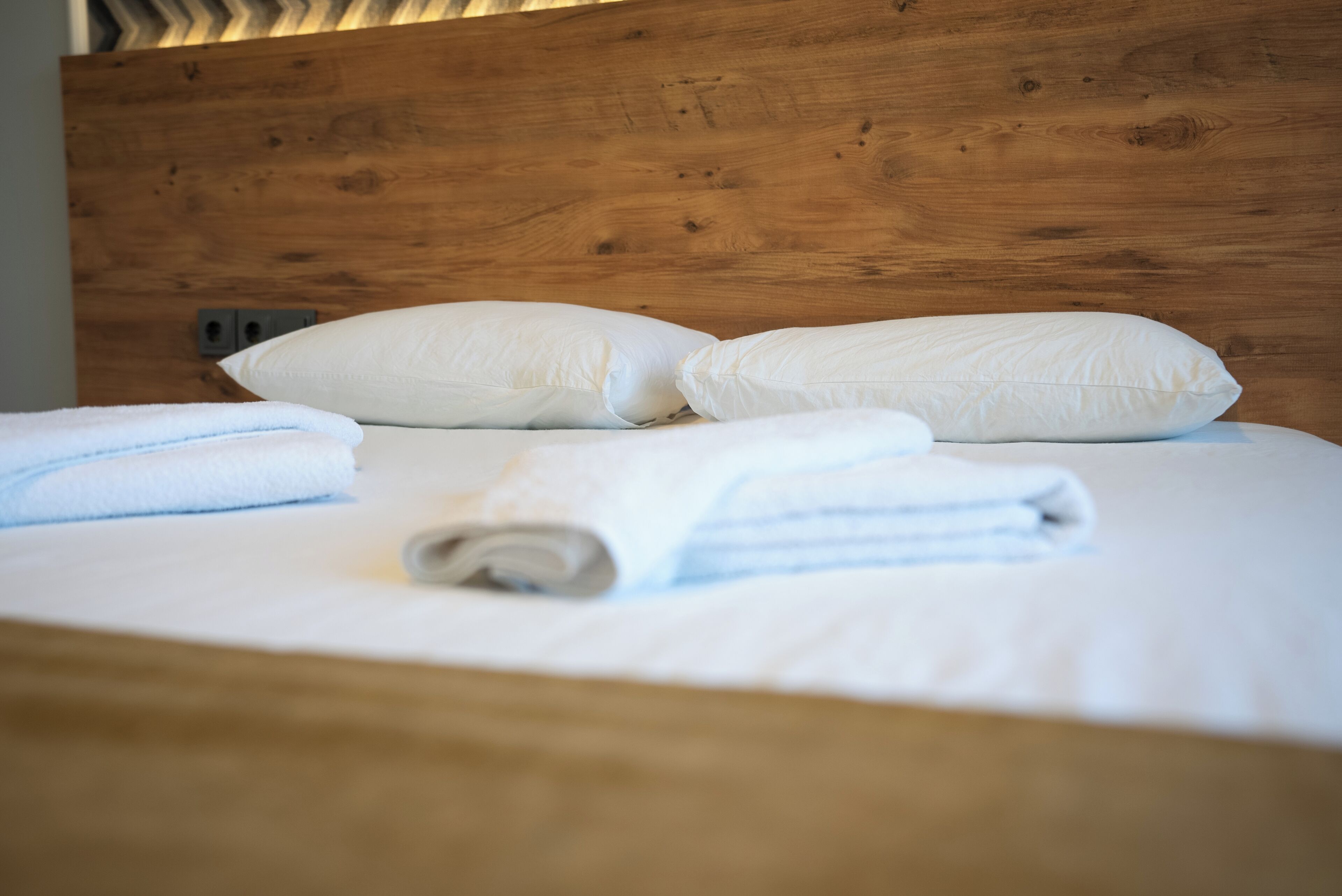 A neatly made bed with white linens and pillows, a wooden headboard, and a visible electrical outlet.