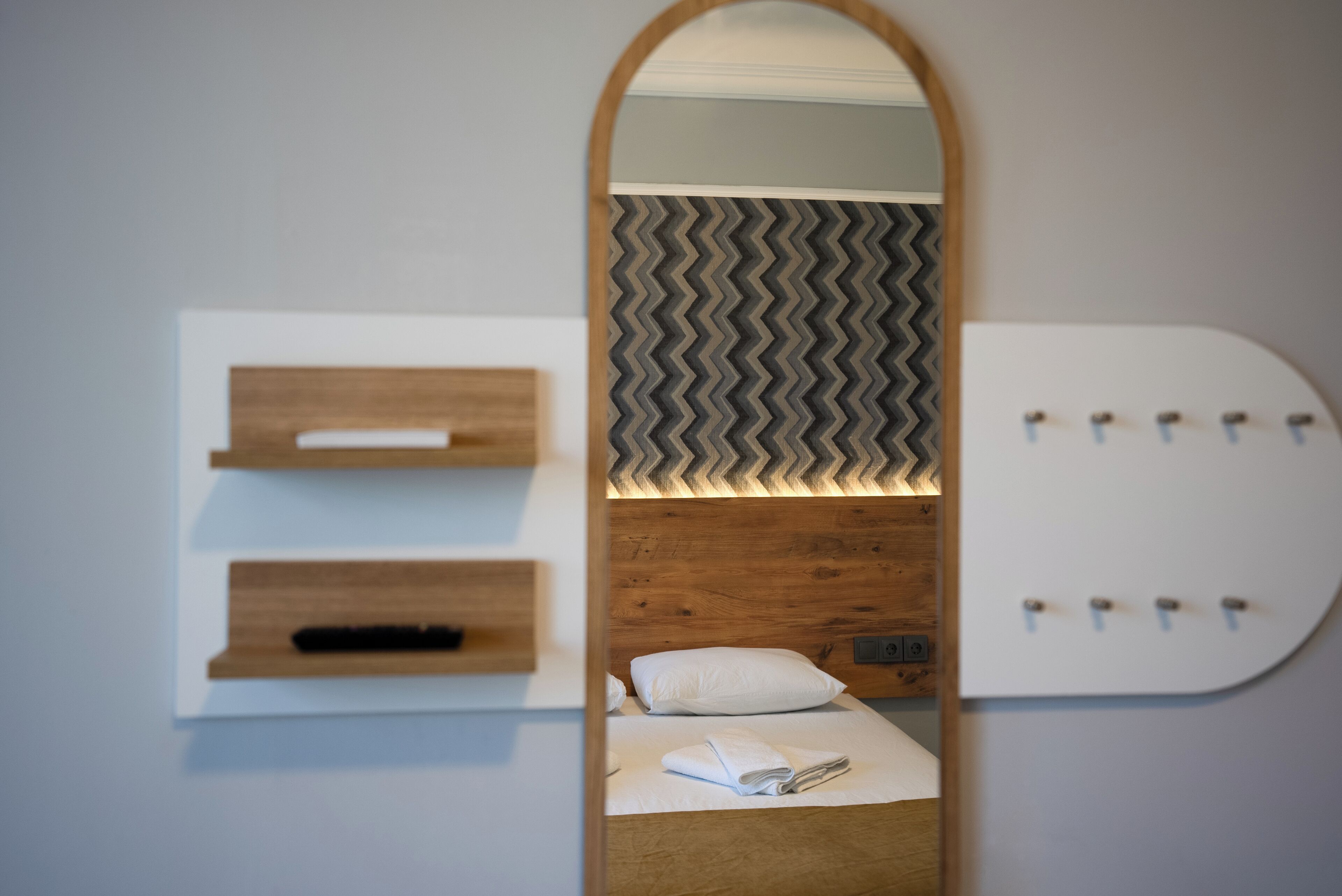 Una habitación de hotel moderna con cabecera de madera, un espejo, una cama con ropa blanca y un estante montado en la pared, además de un papel tapiz decorativo.