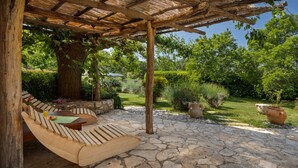 Uma área de estar ao ar livre com bancos de madeira, pérgola, piso de pedra e plantas em vasos.