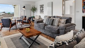 Una sala de estar moderna con un sofá gris, una mesa de centro de madera y vistas al mar a través de un ventanal grande.