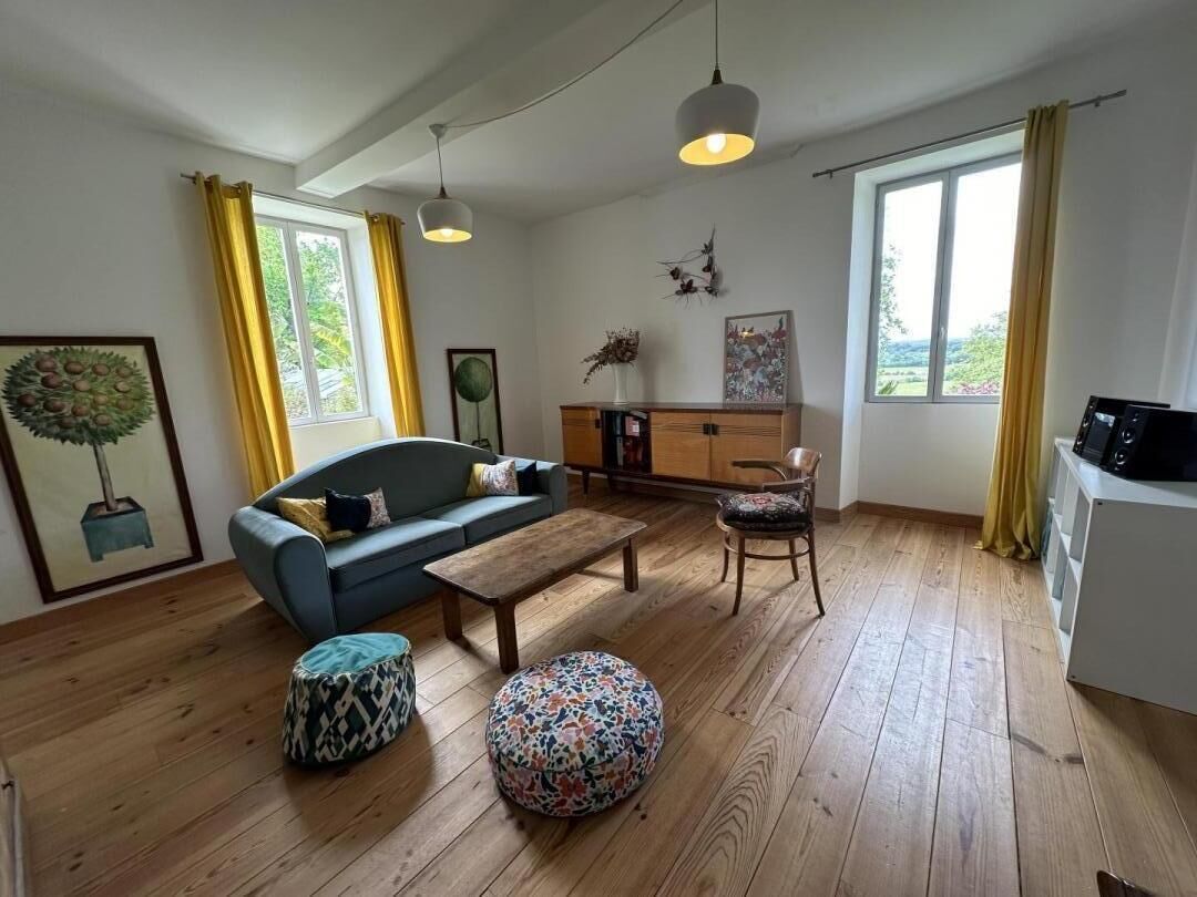 A living room with a sofa, coffee table, and ottoman, wooden flooring, and a view of the outdoors through large windows.