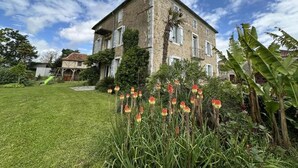 Un bâtiment en pierre à deux étages, avec une pelouse bien entretenue et des plantes en fleurs au premier plan.