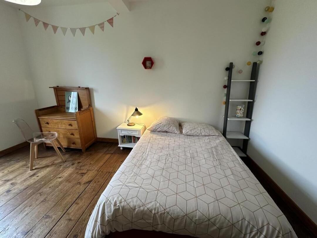 A bedroom with a bed, wooden dresser, chair, bedside table with lamp, and a wall-mounted shelf.