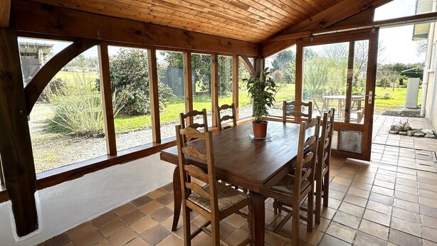 A wooden dining area with a table and chairs, large windows, and a potted plant. - Le Gîte du Sequé - Saint Pandelon (SAINT PANDELON)