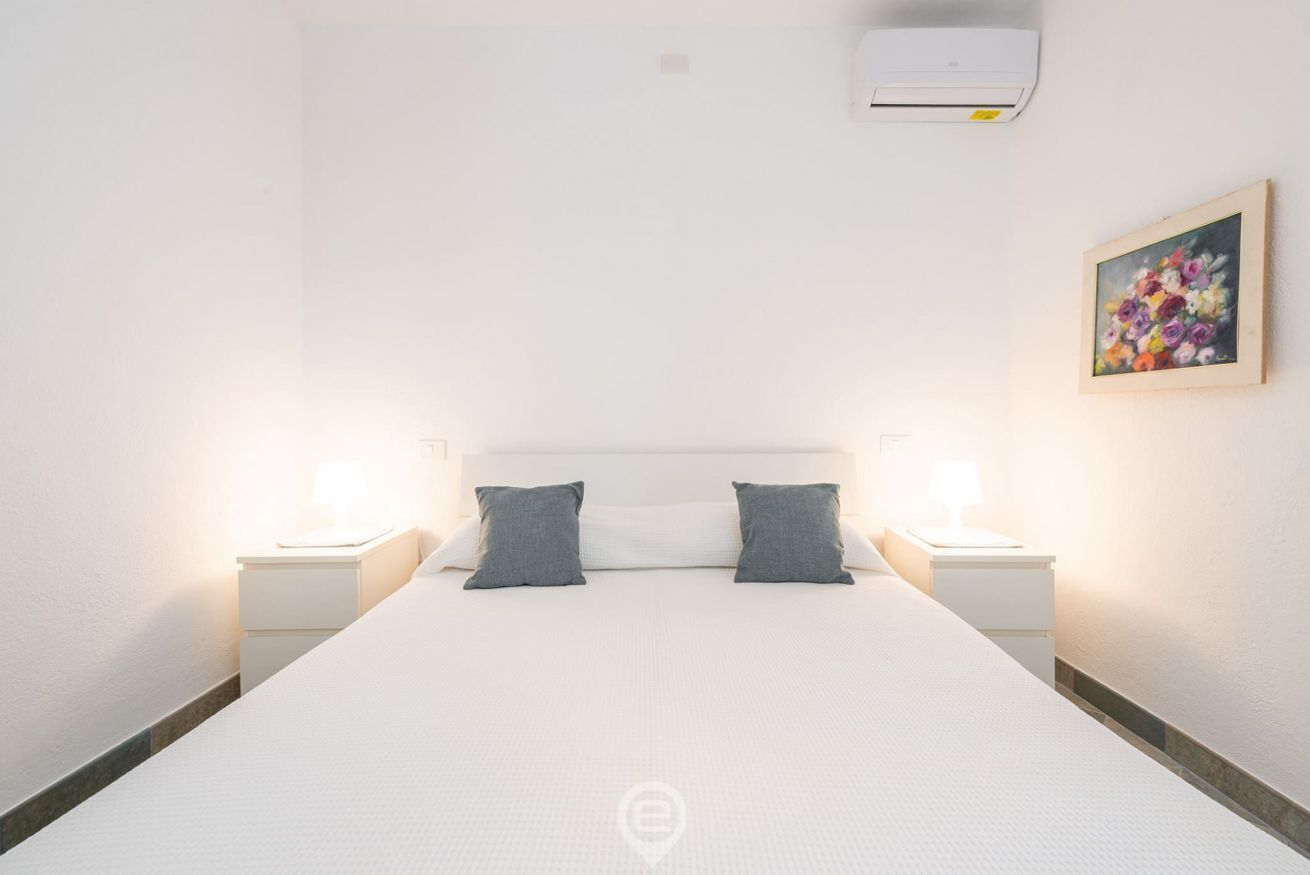 A hotel room with a large bed, two bedside tables with lamps, a wall-mounted air conditioner, and a framed floral painting.