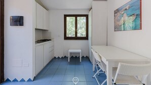 Una cucina compatta con mobili bianchi, un piccolo tavolo bianco, una finestra con vista sul verde e un quadro appeso al muro.