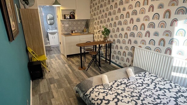 Una habitación compacta con una cama, una mesita pequeña y una silla. Se ve una kitchenette al fondo.