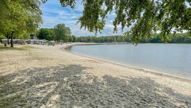 A sandy beach with a calm lake, trees, and a building in the background. - Cozy home in Noordscheschut with WiFi (Noordscheschut)