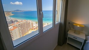 A room with a large window overlooking a beach and ocean, a bedside table with a lamp, and a wall socket. - Seaside penthouse apartment with sea views and swimming pool (Calpe)
