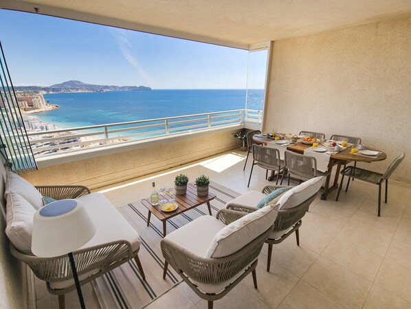 A balcony with wicker furniture, a dining table set for breakfast, and a view of the sea and cityscape.