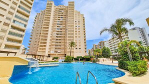 A swimming pool surrounded by high-rise buildings, with palm trees and a clear sky. - Seaside penthouse apartment with sea views and swimming pool (Calpe)