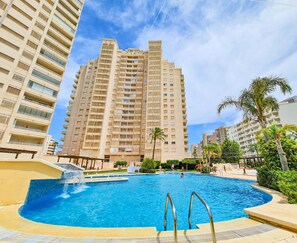A swimming pool surrounded by high-rise buildings, with palm trees and a clear sky.