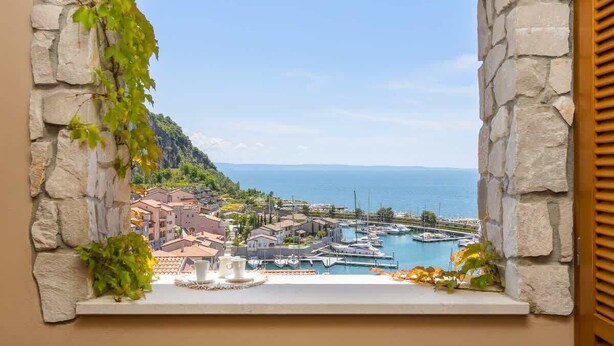 A stone-framed window with a view of a coastal town, a marina, and the sea.