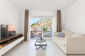 A modern living room with a flat-screen TV, a sofa, a coffee table, and a balcony with a view of buildings. - La tempesta perfetta - Portopiccolo Apartments (Duino-Aurisina)