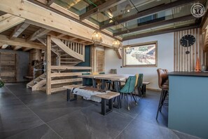 Amplia habitación con escalera de madera, zona de comedor con mesa y sillas, barra y un gran ventanal con vistas.