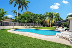 Un patio trasero con piscina, rodeado por una cerca de madera y un área de patio techada con sillones de descanso.