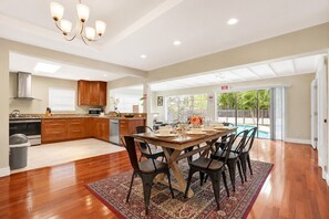 Un comedor amplio con una mesa y sillas de madera, una cocina al fondo y vista a una piscina a través de puertas de vidrio grandes.