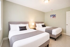 A hotel room with two beds, each with a grey headboard and white bedding. There is a nightstand with a lamp between the beds, and a door to the right.