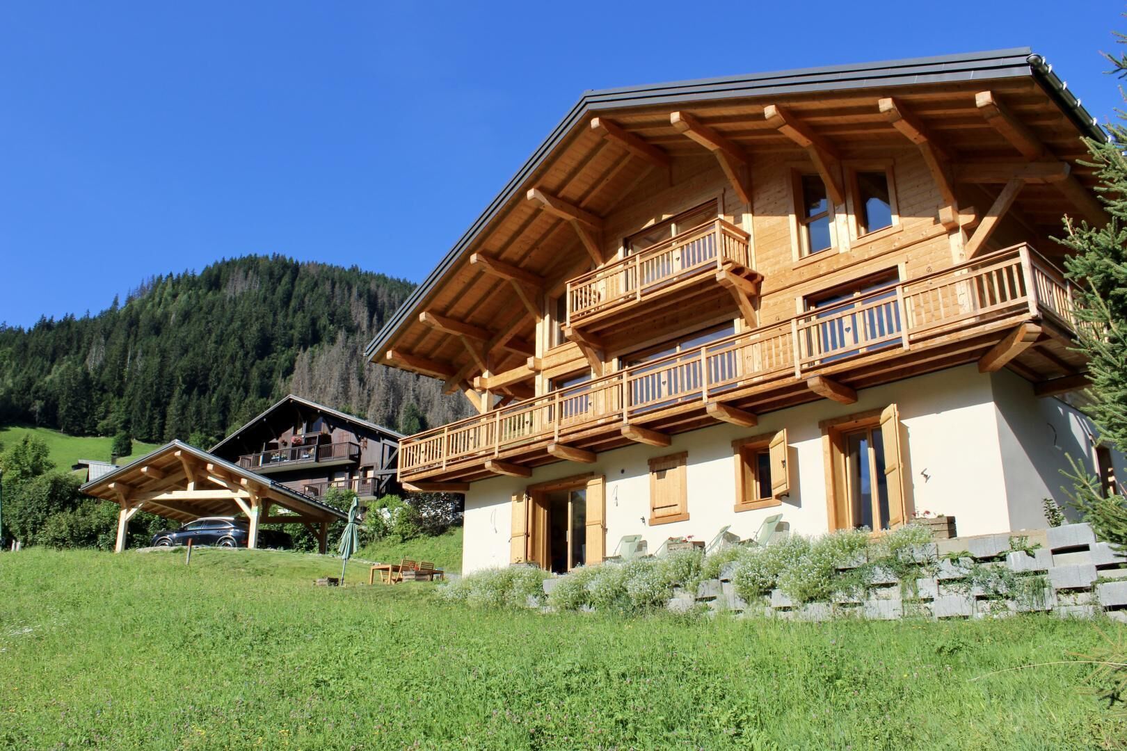 Een houten chalet met twee verdiepingen en balkons, omgeven door een grasveld en een bebost berglandschap.