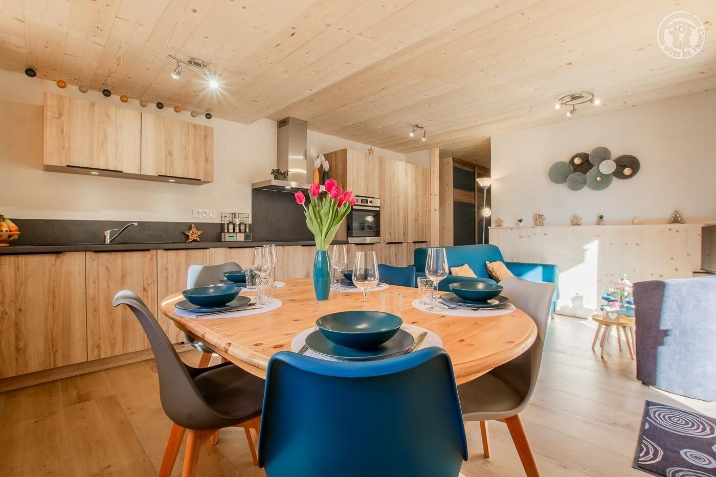 Une cuisine moderne avec des meubles en bois, une table à manger entourée de chaises bleues et un vase rempli de fleurs rouges.