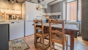 A compact kitchen with modern appliances, a dining area with wooden chairs, and a view of a wooden deck through a window. - LA P'tite Pierrue - Arvillard (ARVILLARD)