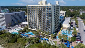 Un hotel moderno de gran altura con varios balcones, un área de piscina con sombrillas y un estacionamiento.