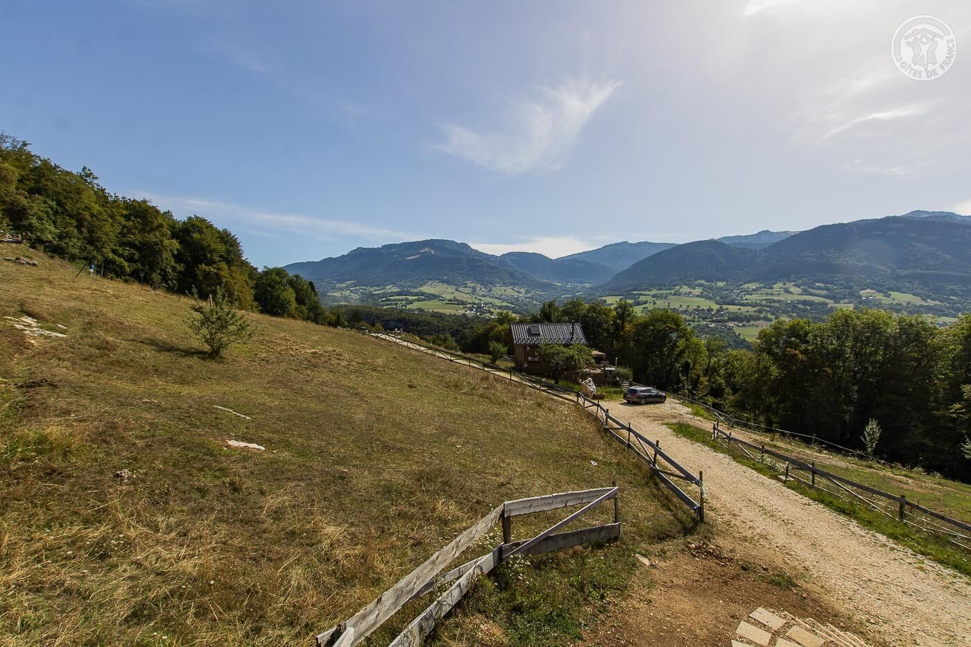 Paisaje rural con un camino de tierra que conduce a un pequeño edificio con techo de metal, rodeado de árboles y montañas al fondo.
